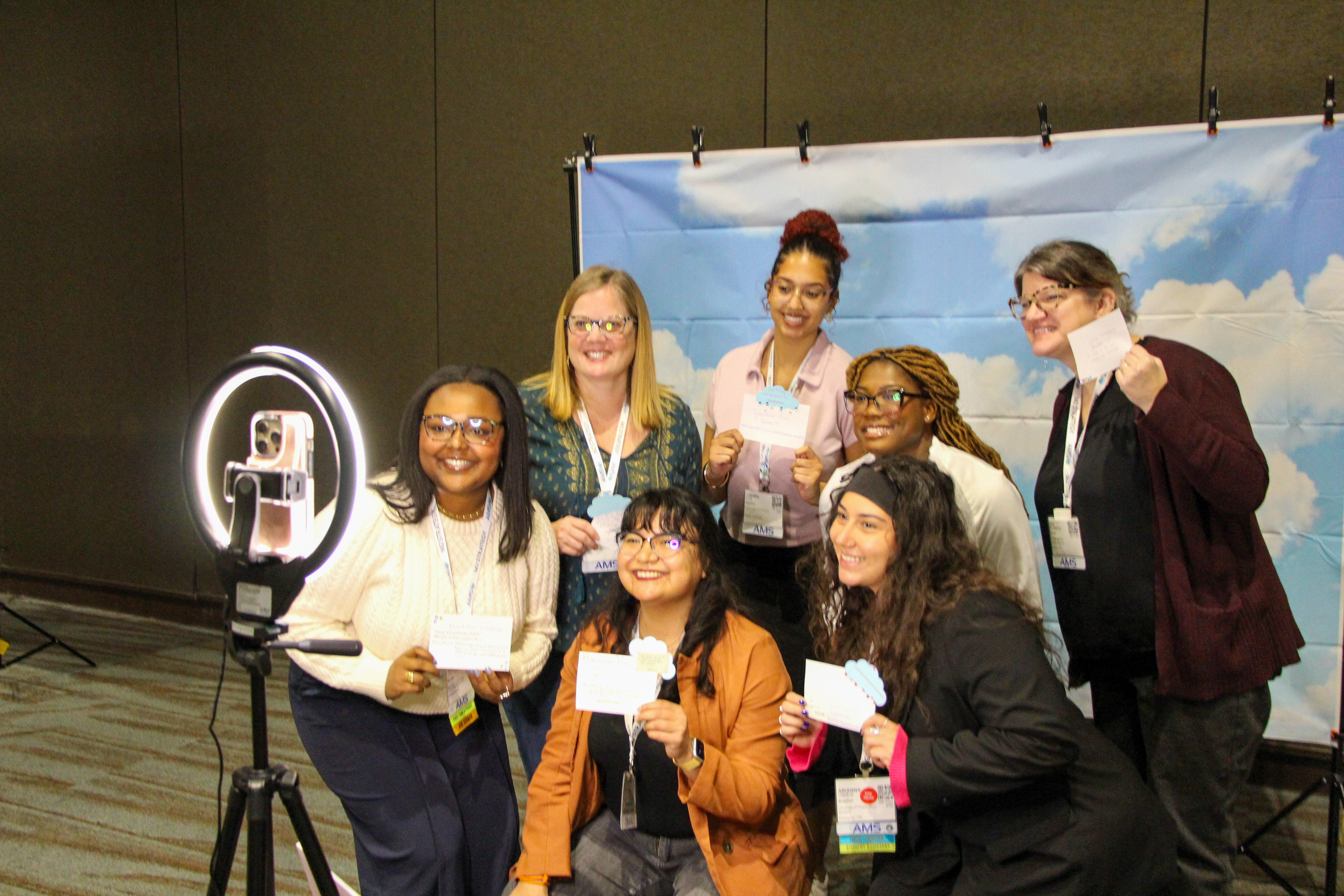 A group of seven people smiling at a camera with a selfie ring light. They hold small cards, standing before a backdrop featuring blue skies with clouds. The scene conveys positivity and camaraderie.