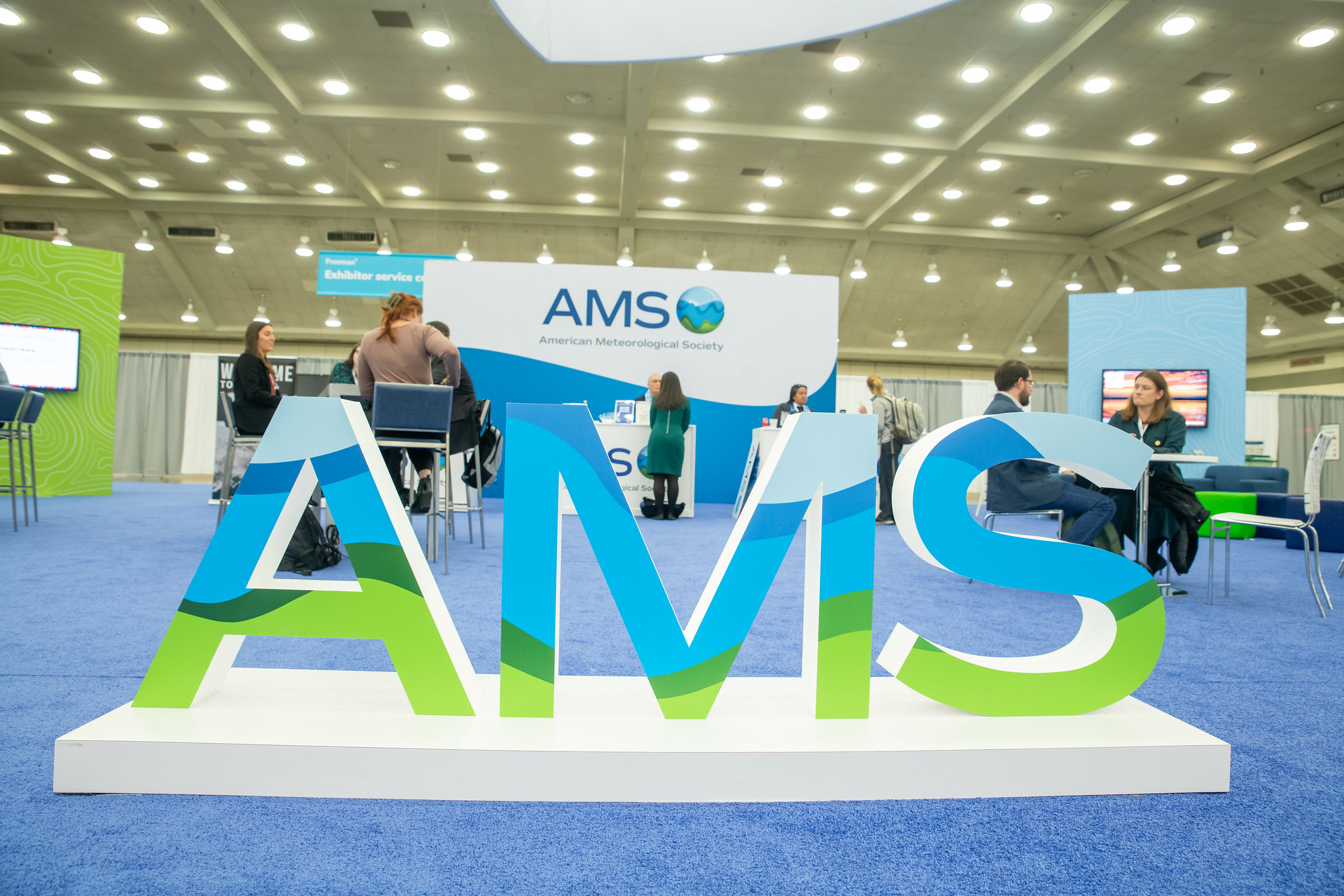Large "AMS" letters in a convention center with blue carpet and bright lighting. People are gathered around a booth for networking and discussions.