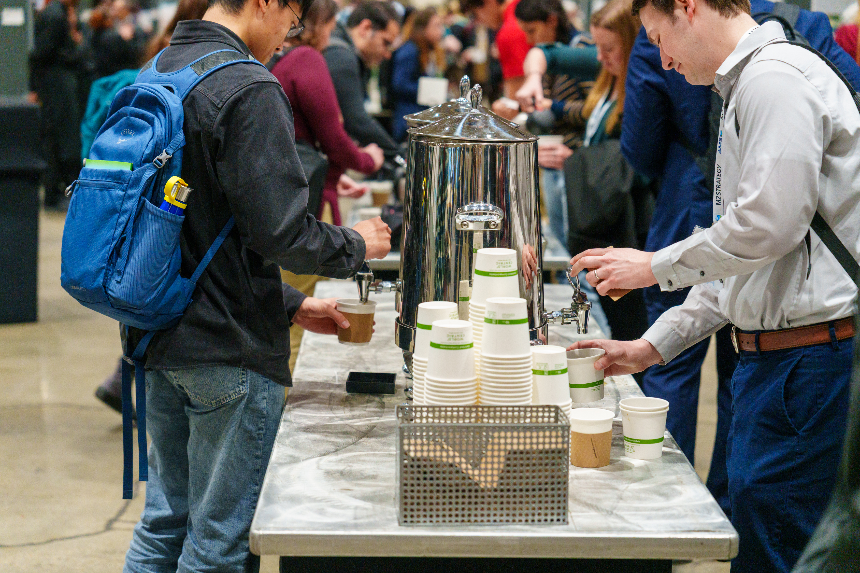  People with backpacks gather around a coffee station at an event, serving themselves from a large metal urn. Stacks of paper cups are on the counter. The scene is busy and social.