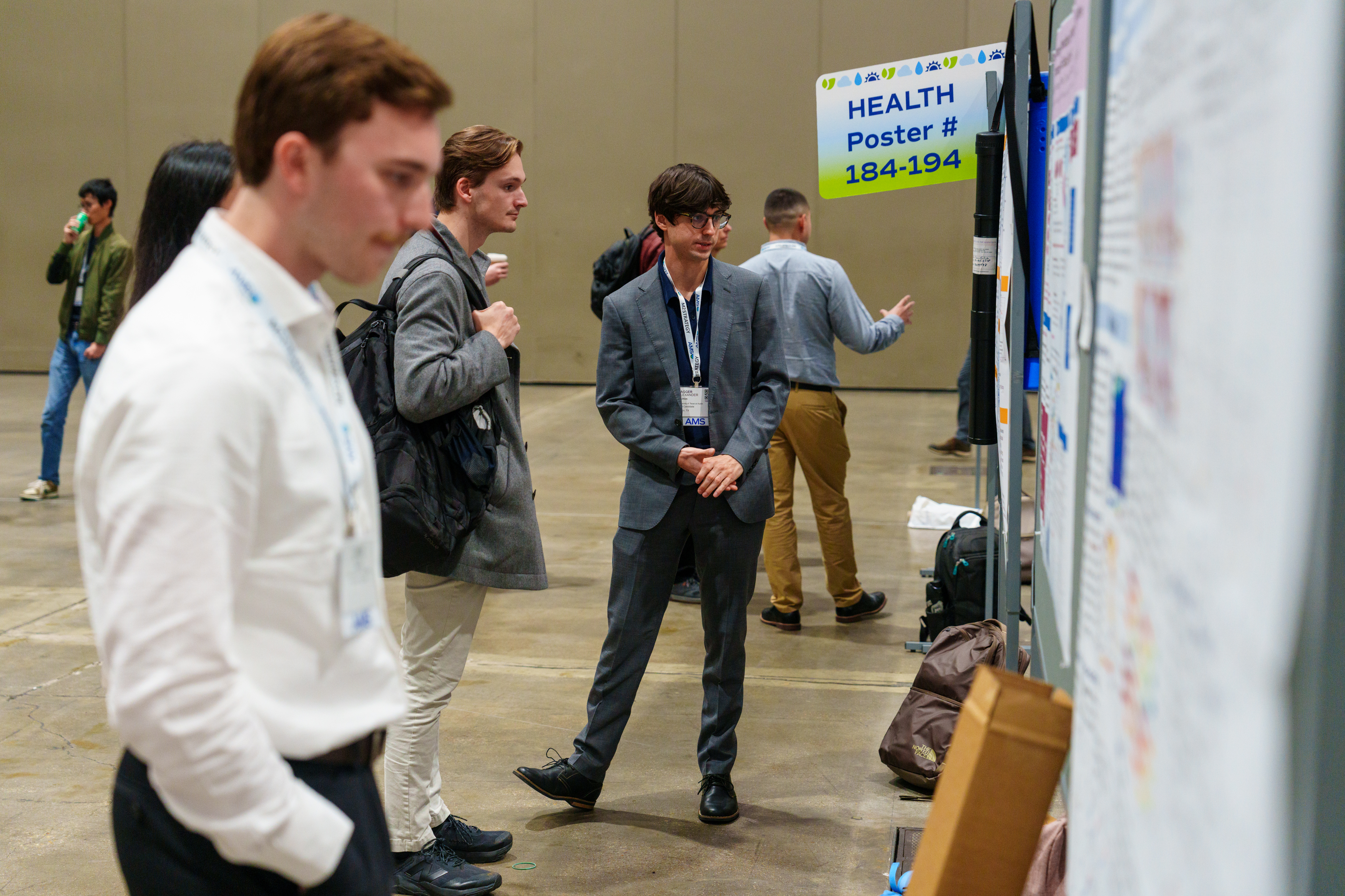 A group of attendees in business attire engage in discussion at a conference poster session. A sign reads "HEALTH Poster #184-194.