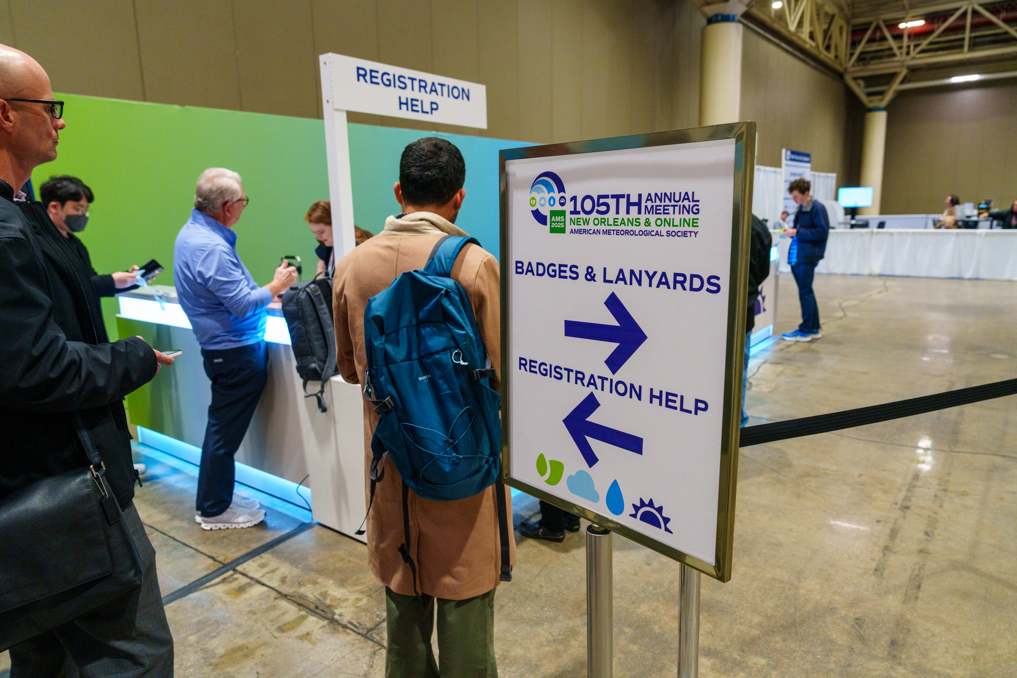 People stand in a line at the conference registration help desk. In the foreground of the image, there is a large sign for the 105th Annual Meeting. The sign directs attendees with large dark blue arrows to the right for badges and lanyards or to the left for the Registration Help desk.