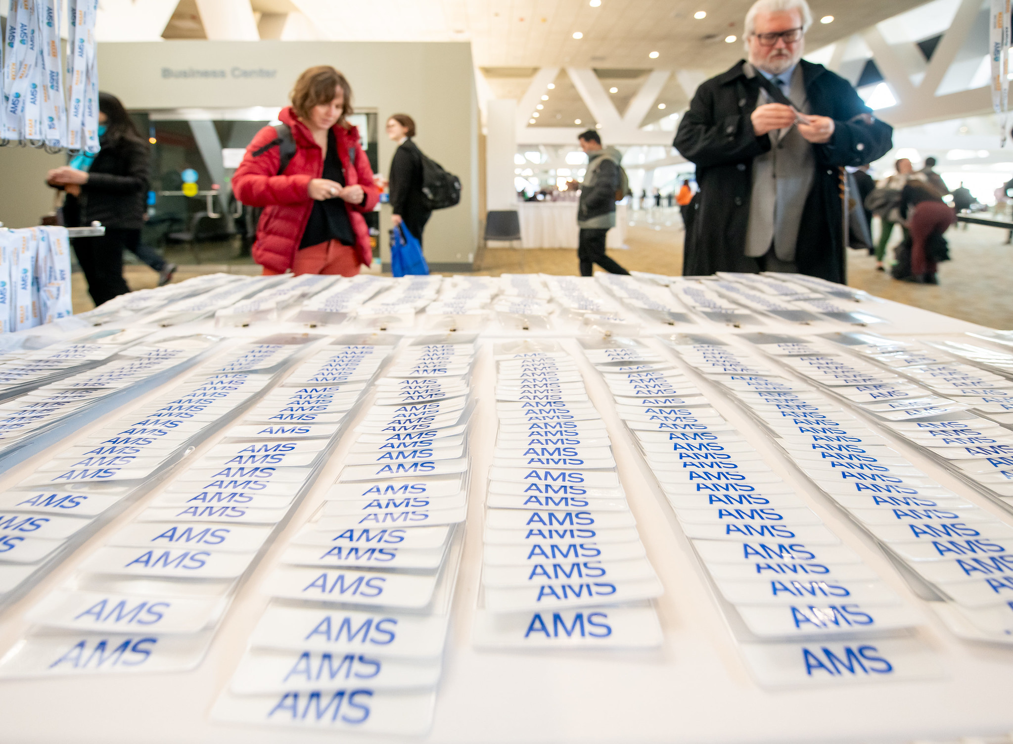 A table in the foreground of the image is filled with many rows of white badge holders labeled "AMS" in blue at a conference venue. Two people in the background are picking up badges and attaching them their lanyard.
