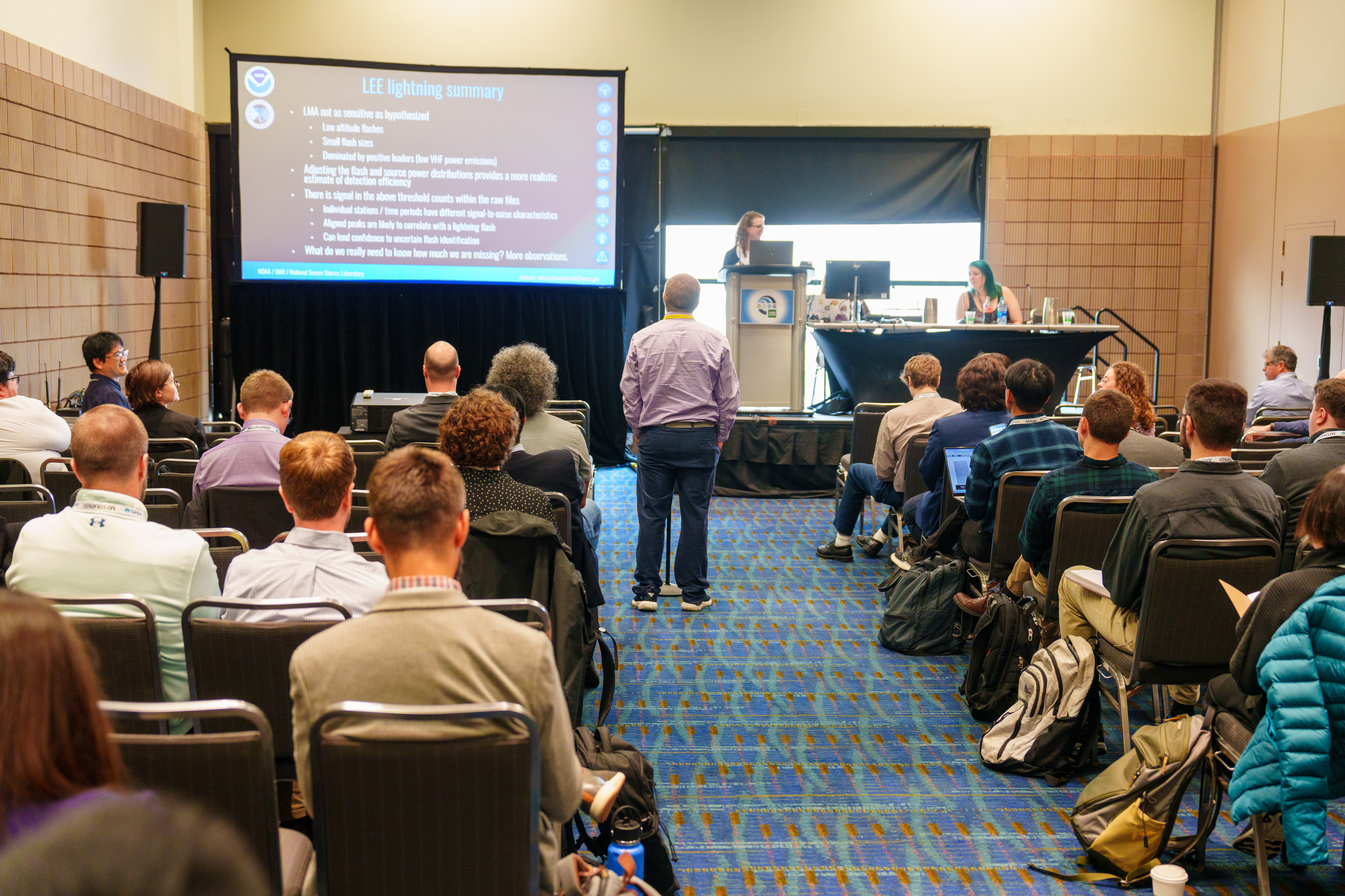 A conference room filled with seated attendees facing away from the camera and towards a speaker at a podium. An attendee is standing in the aisle near the stage to ask the presenter at the podium a question. A presentation screen on the upper left side of the image displays slides