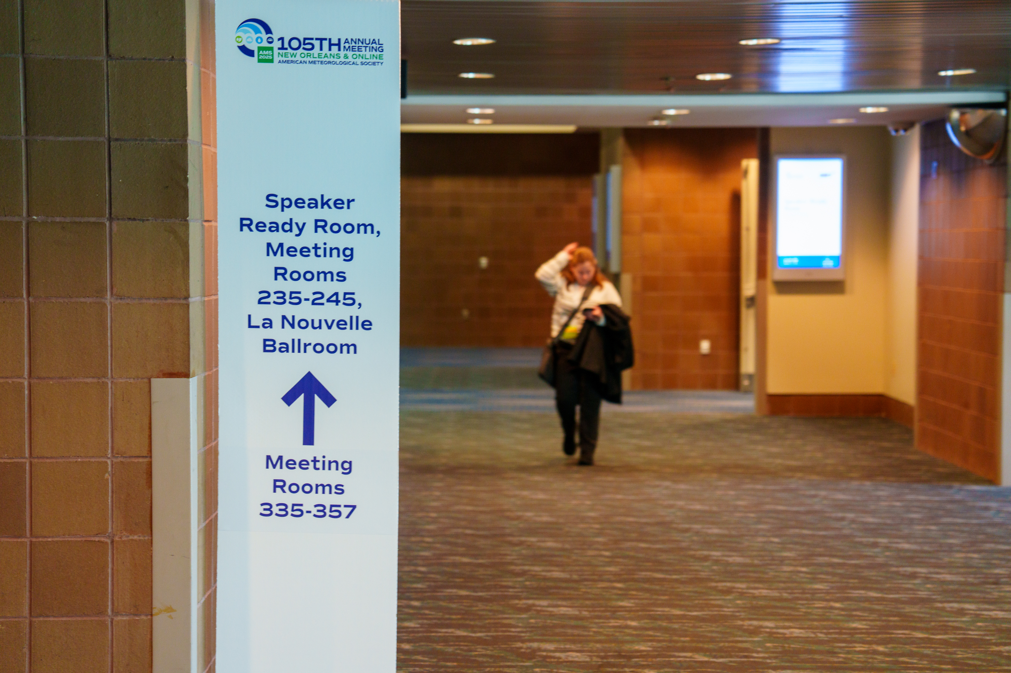 Convention center hallway with directional sign for speaker ready rooms, La Nouvelle Ballroom, and meeting rooms; attendee walks in background.