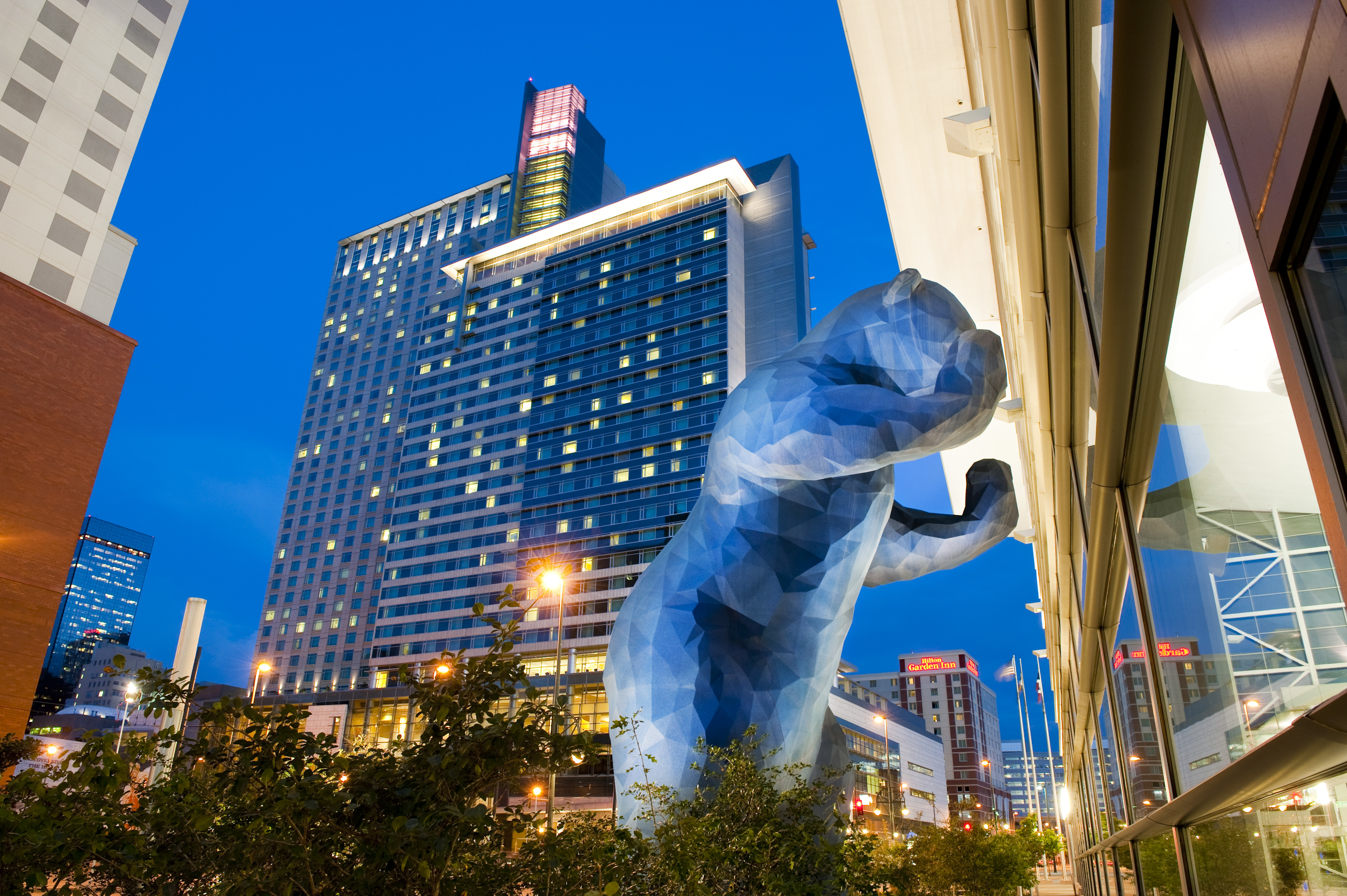 Giant blue bear sculpture peeking into the Colorado Convention Center, with the illuminated Hyatt Regency Denver towering in the background at dusk.