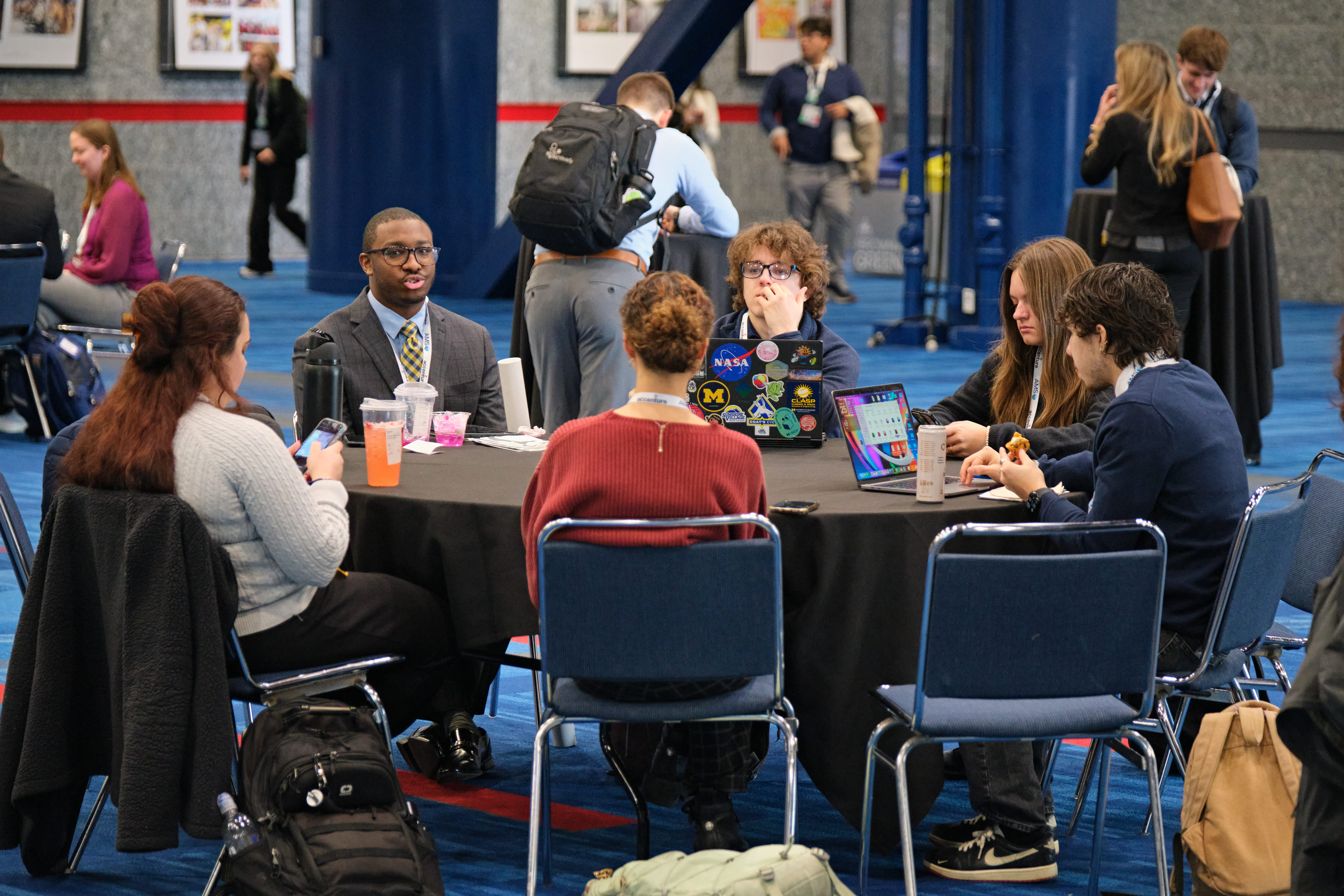 Attendees sit around a conference table, talking over laptops, drinks, and notebooks in a busy hall; the mood is engaged and collaborative.