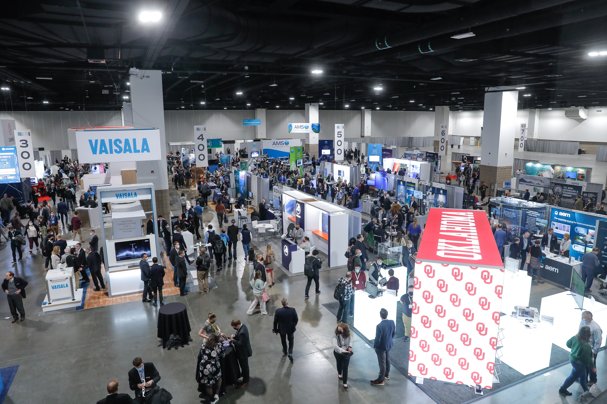 A bustling exhibit hall with numerous rows of booths displaying technology and products. People are mingling and networking in the aisles under bright lights.