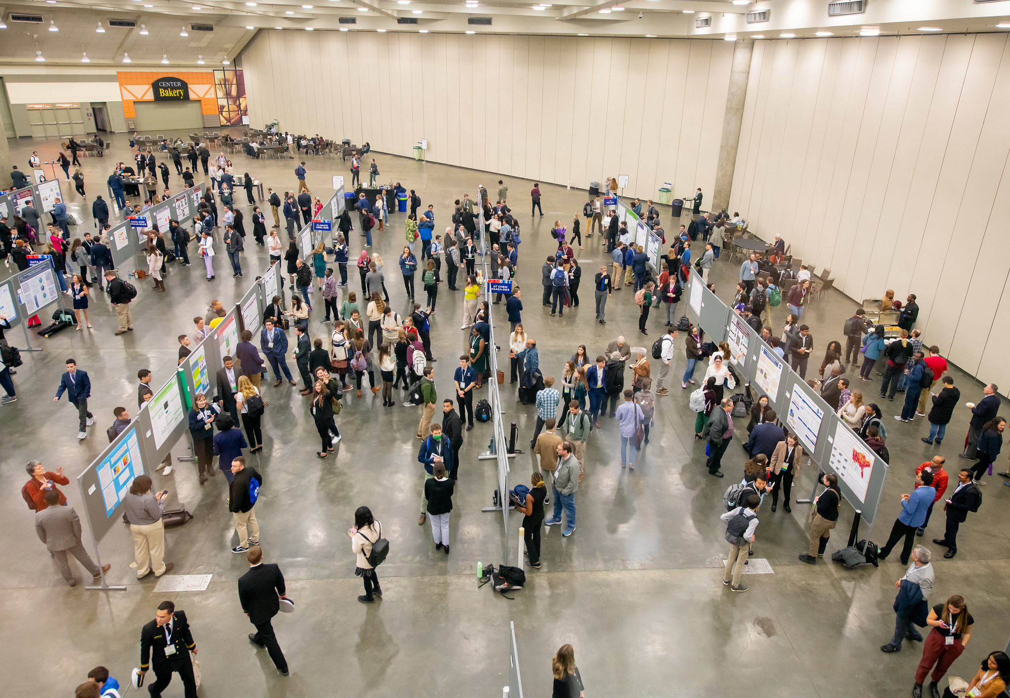 A large conference hall filled with attendees examining and discussing poster presentations displayed on panels.