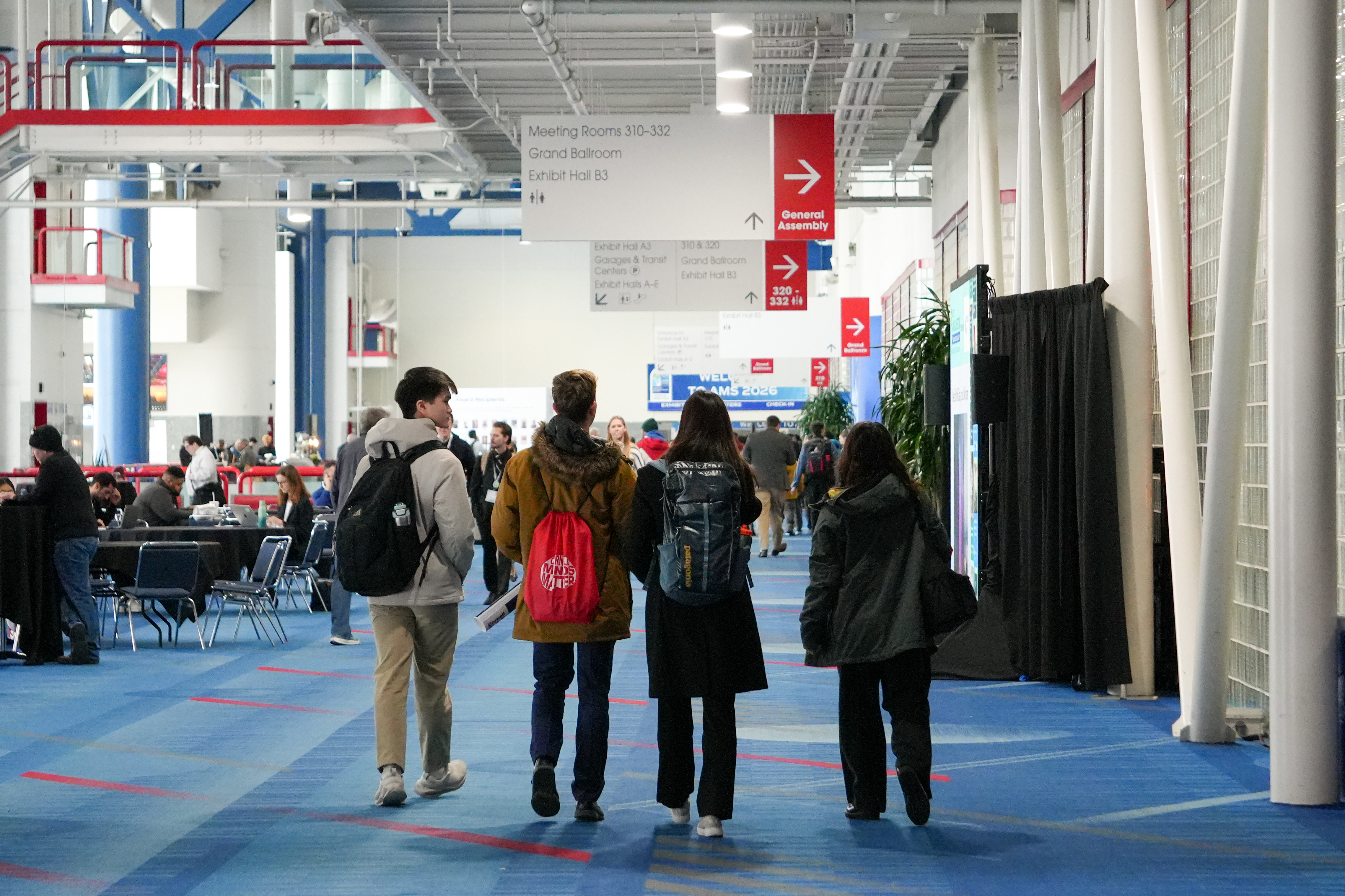 Visitors walk through a bright convention center, following red directional signs past seating areas and booths beneath an industrial ceiling.