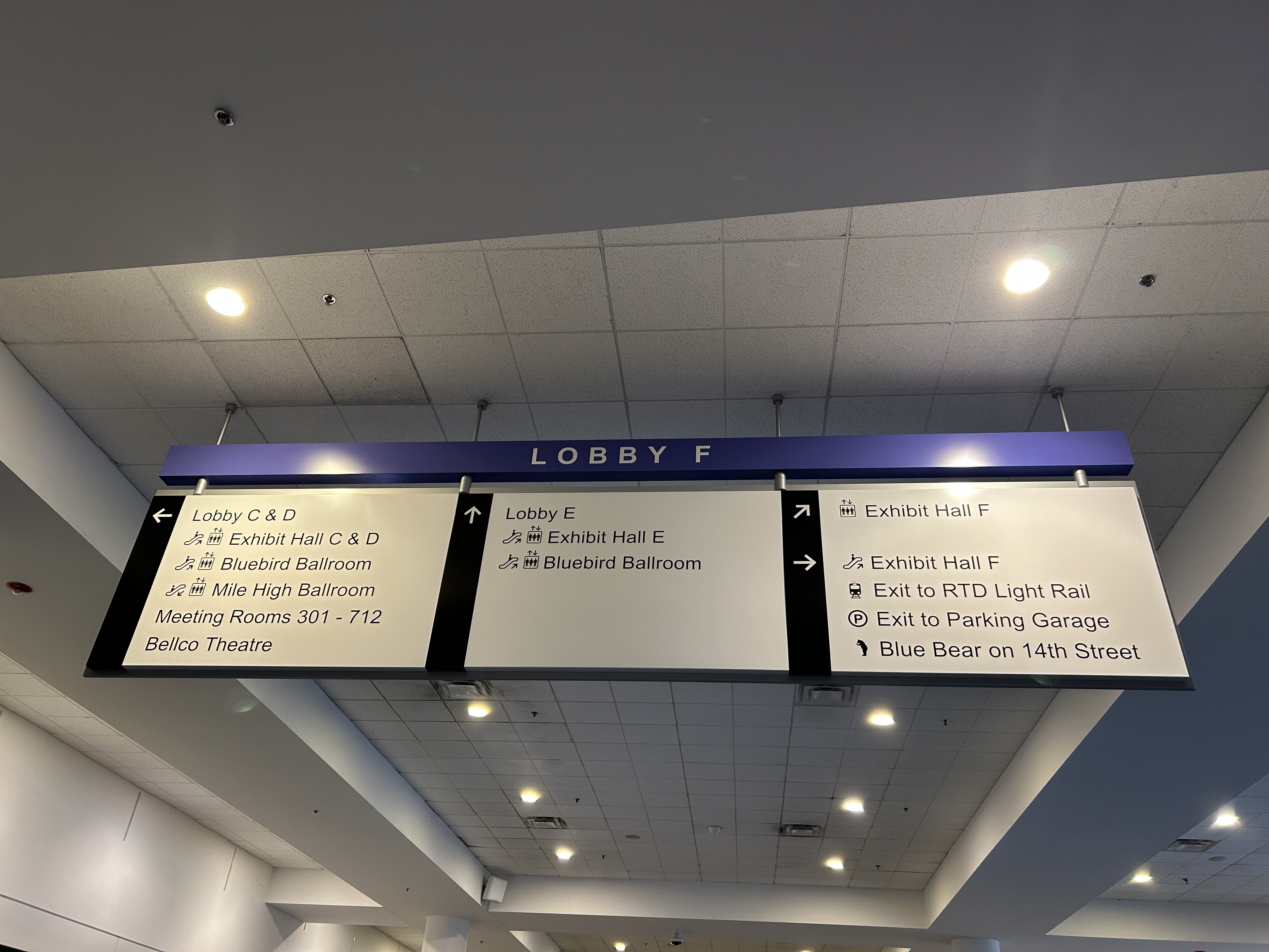 Overhead convention-center sign labeled “Lobby F” lists directions to exhibit halls, ballroom, parking garage, light rail, and nearby theater.