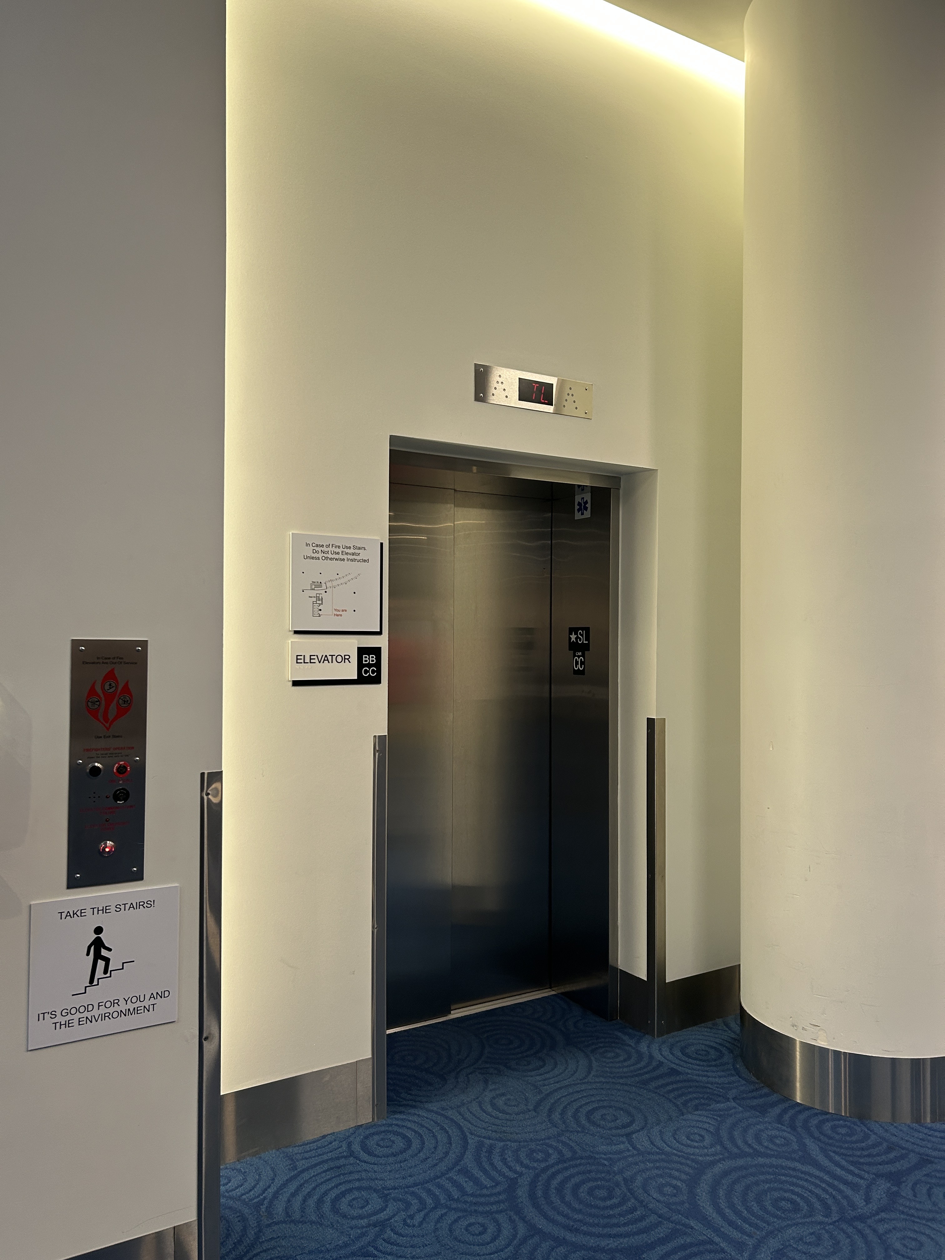 Modern convention center elevator lobby with stainless-steel doors, call buttons, accessibility signage, curved white walls, warm lighting, and blue patterned carpet.
