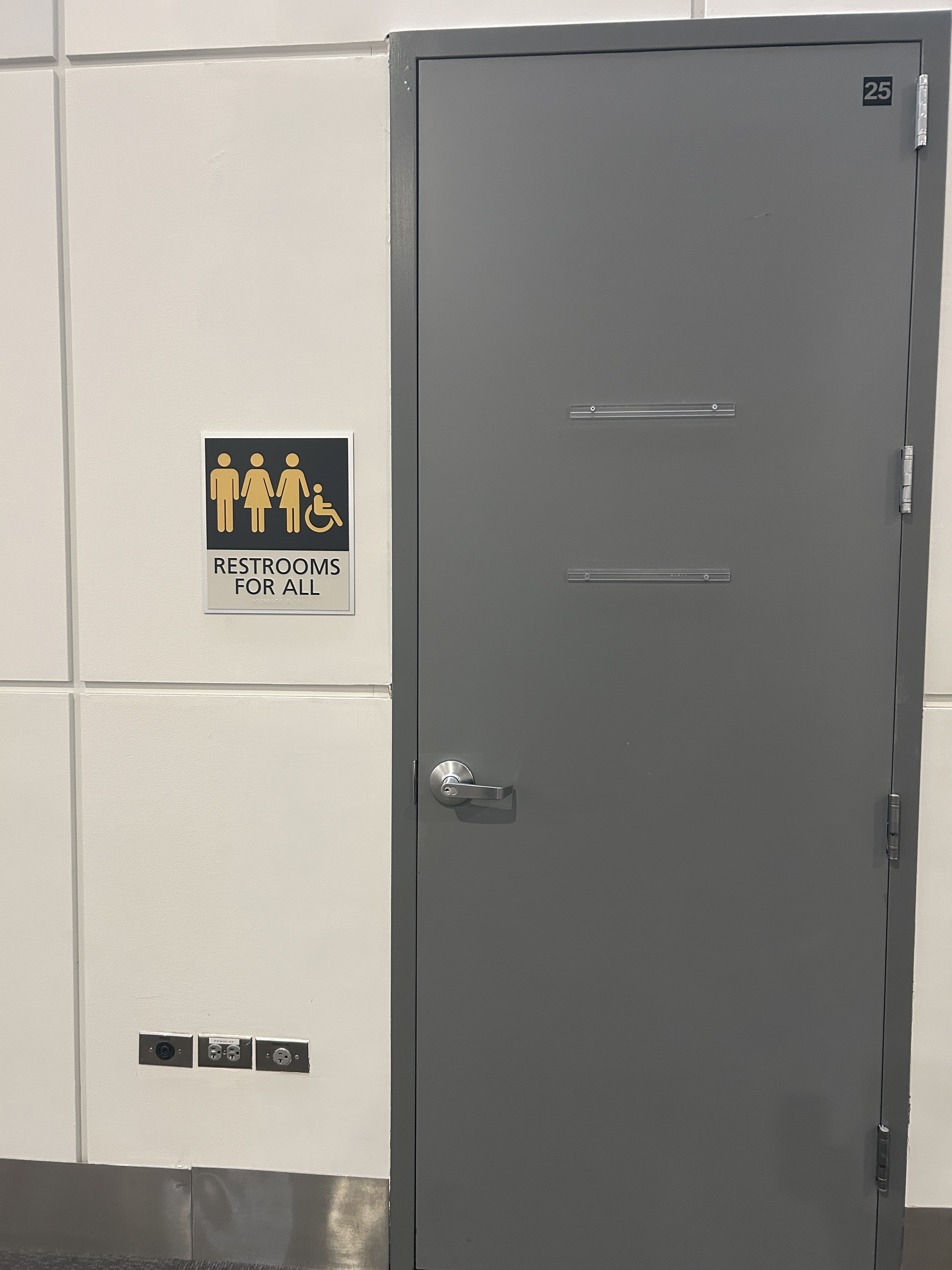 Image shows a closed, gray door beside a dark navy “RESTROOMS FOR ALL” sign with three gold people icons and a wheelchair icons mounted on a white tiled hallway wall.